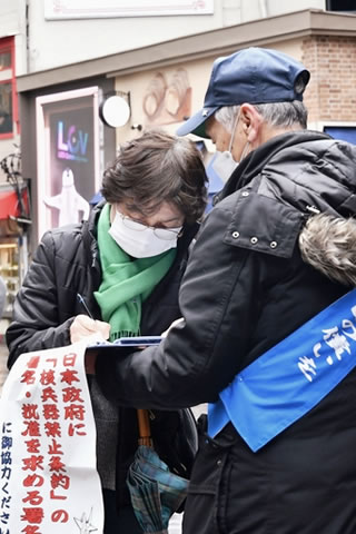 署名板を支え持つ東友会のタスキをかけた被爆者と、署名をする通行人。